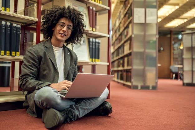 A Brazilian student using a laptop at a modern university library in the USA, surrounded by books and other students. The scene represents academic pursuit and the resources available to international students.